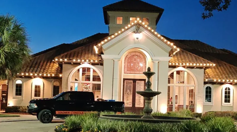 Clubhouse at dusk with warm-white bulbs along every roofline and gable, a palm tree, a stone fountain and a black Wagner Consolidated pickup truck parked out front